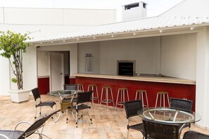 Um pátio externo com mesa de vidro, cadeiras de vime, um revestimento de azulejos vermelhos e um vaso com planta.