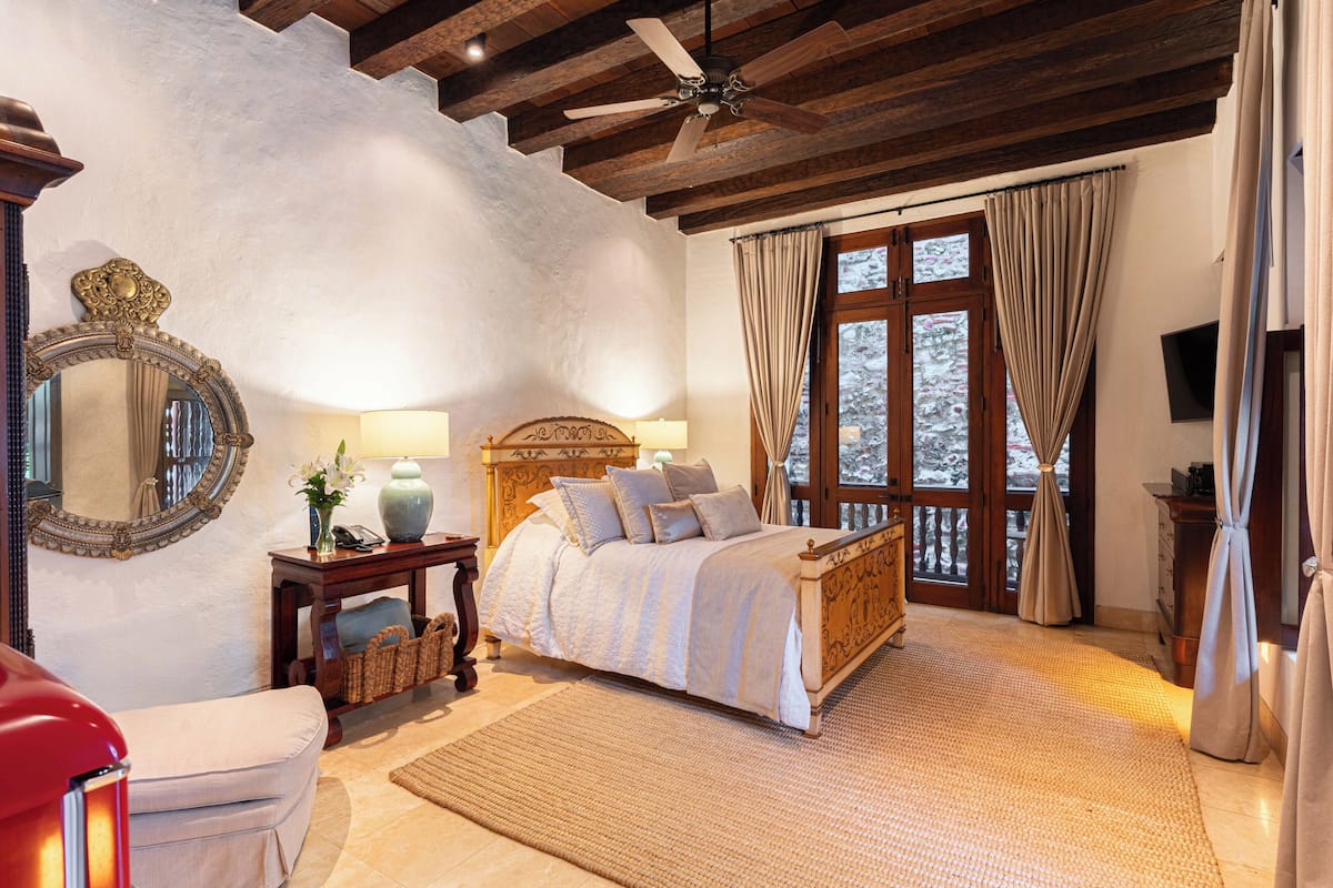 Une chambre à coucher avec un lit, une tête de lit en bois, un ventilateur de plafond, un grand miroir, une table de chevet, une lampe, une fenêtre avec des rideaux et une vue sur les arbres à l’extérieur.