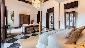 A hotel room with a checkered floor, a large bed with patterned pillows, a chandelier, wooden furniture, and a view through an open door. - CASA ESTRADA LUXURY VILLA (Cartagena)