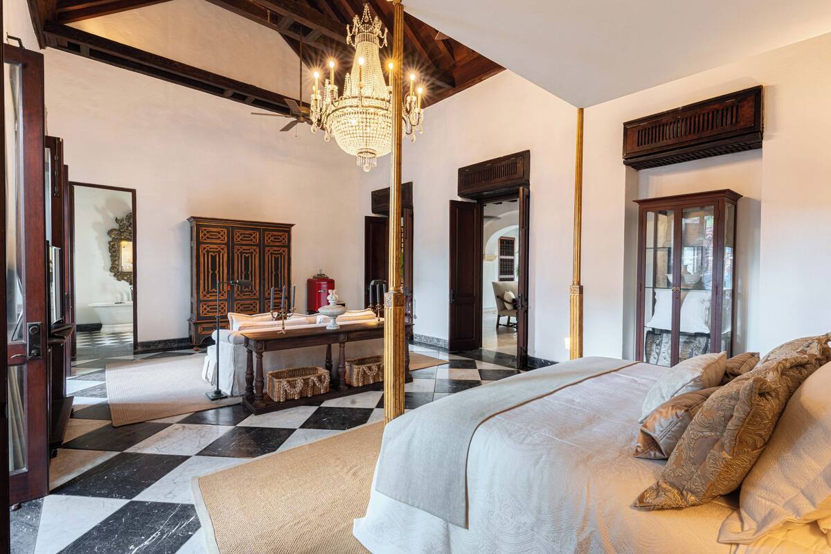 Une chambre d’hôtel avec un sol à carreaux, un grand lit agrémenté d’oreillers à motifs, un lustre, des meubles en bois et une vue sur l’extérieur, visible à travers une porte ouverte.