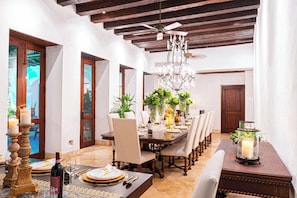 Uma sala de jantar com mesa de madeira posta para refeição, rodeada por cadeiras, e um candelabro acima.