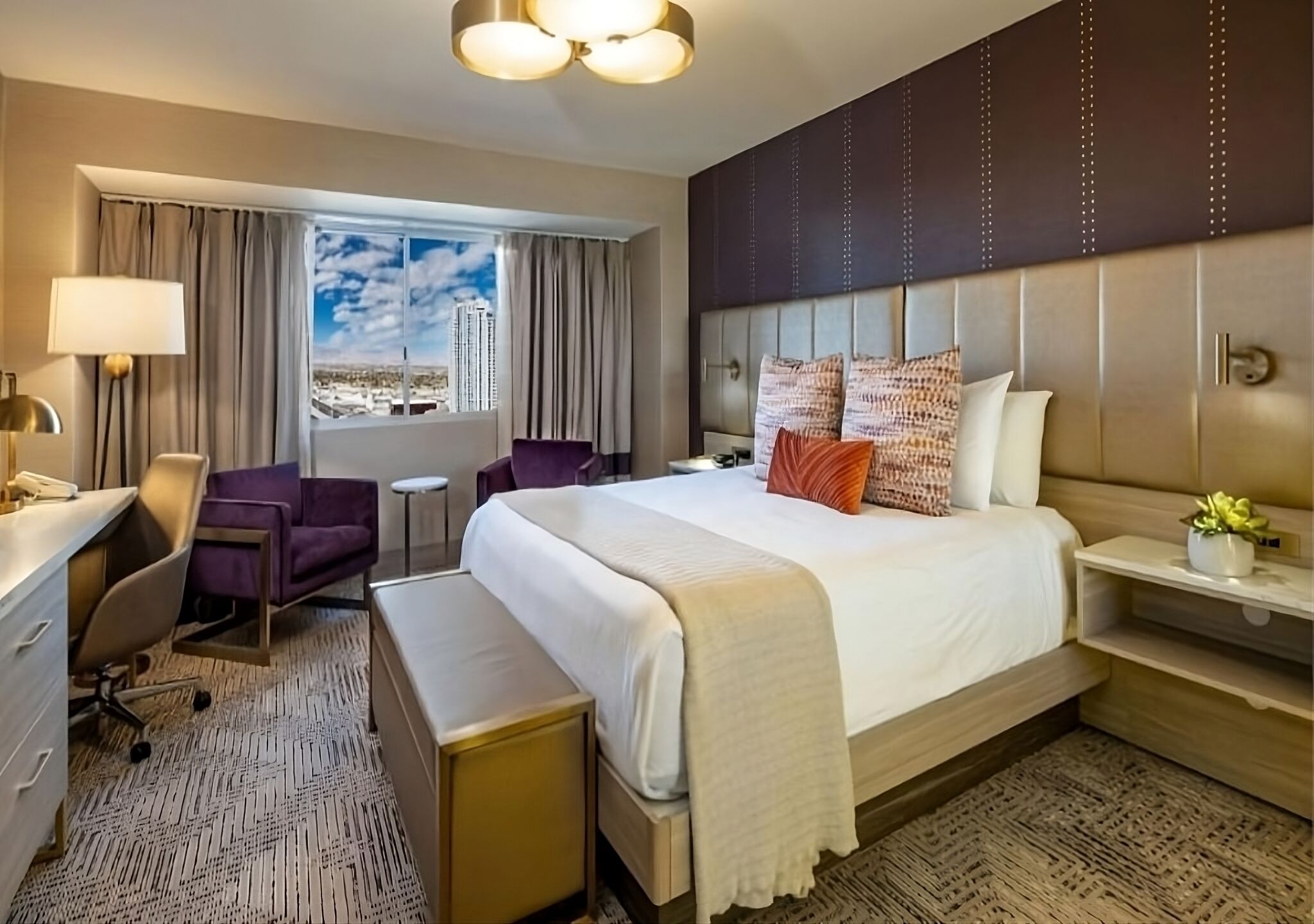 A hotel room with a bed, desk, chair, and a view of the cityscape.