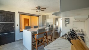 Una cocina moderna con una isla central, taburetes de bar, ventilador de techo y encimera de mármol.