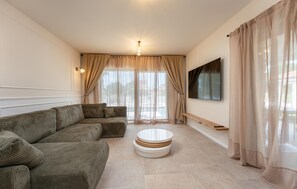 Ein Wohnzimmer mit einem Ecksofa, einem runden Couchtisch, einem flachbildfernseher an der Wand und einem großen Fenster mit durchsichtigen Vorhängen.