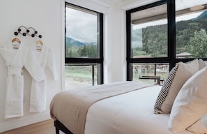 A bedroom with a bed, a window with a view of mountains, and two white robes hanging on the wall. - The Corner Stoke by Revelstoke Vacations (Revelstoke)