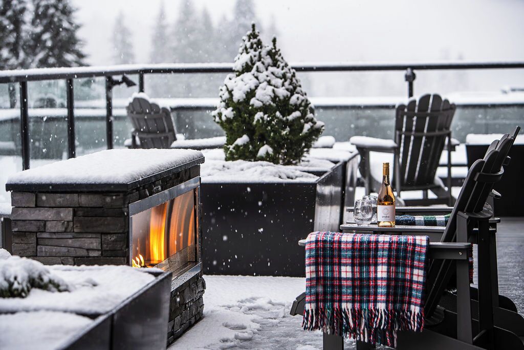 A snow-covered outdoor seating area with a stone fireplace, a Christmas tree, and a table with a bottle and glasses.
