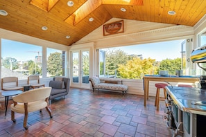 Une véranda avec un plafond en bois, de grandes fenêtres, un canapé, des fauteuils, une table et un poêle.