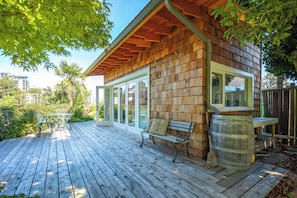 Une terrasse en bois, dotée de bancs et d’un grand tonneau, entourée de verdure et bordée d’un bâtiment aux grandes fenêtres.