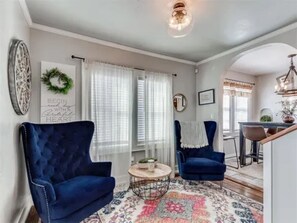 Ein Wohnzimmer mit zwei blauen Sesseln, einem runden Couchtisch, einem floralen Teppich und einem Essbereich im Hintergrund.