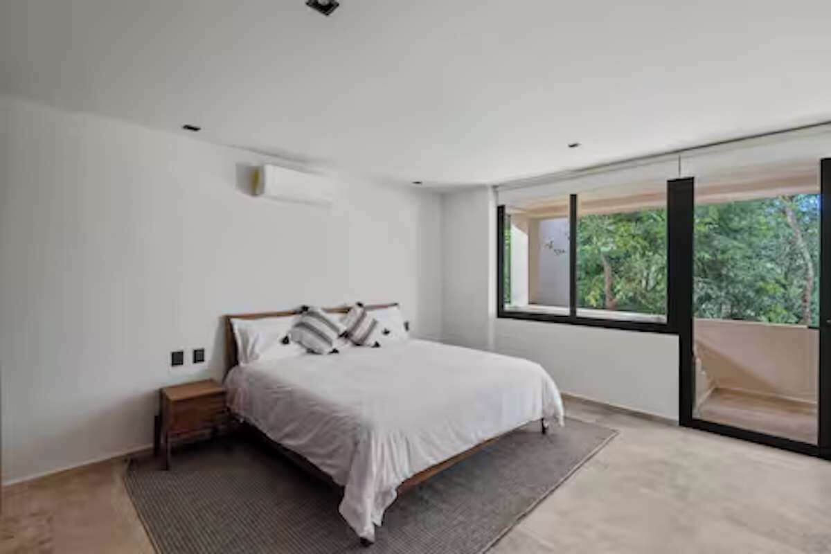 A modern bedroom with a bed, a small bedside table, a large window, and a view of greenery outside.