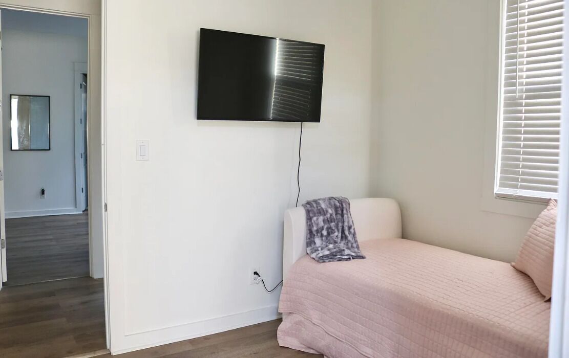 A bedroom with a bed, a TV mounted on the wall, and a window with blinds.