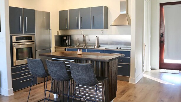 A modern kitchen with dark cabinets, stainless steel appliances, a central island with bar stools, and a large window. - Welcome to 30A Glass House — a bright, modern coastal retreat (Santa Rosa Beach)