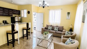 A living room with a sofa, chairs, a coffee table, and a kitchen area with cabinets and appliances. - 2BR Apartment with Ocean & Mountain Views (Puerto Plata)