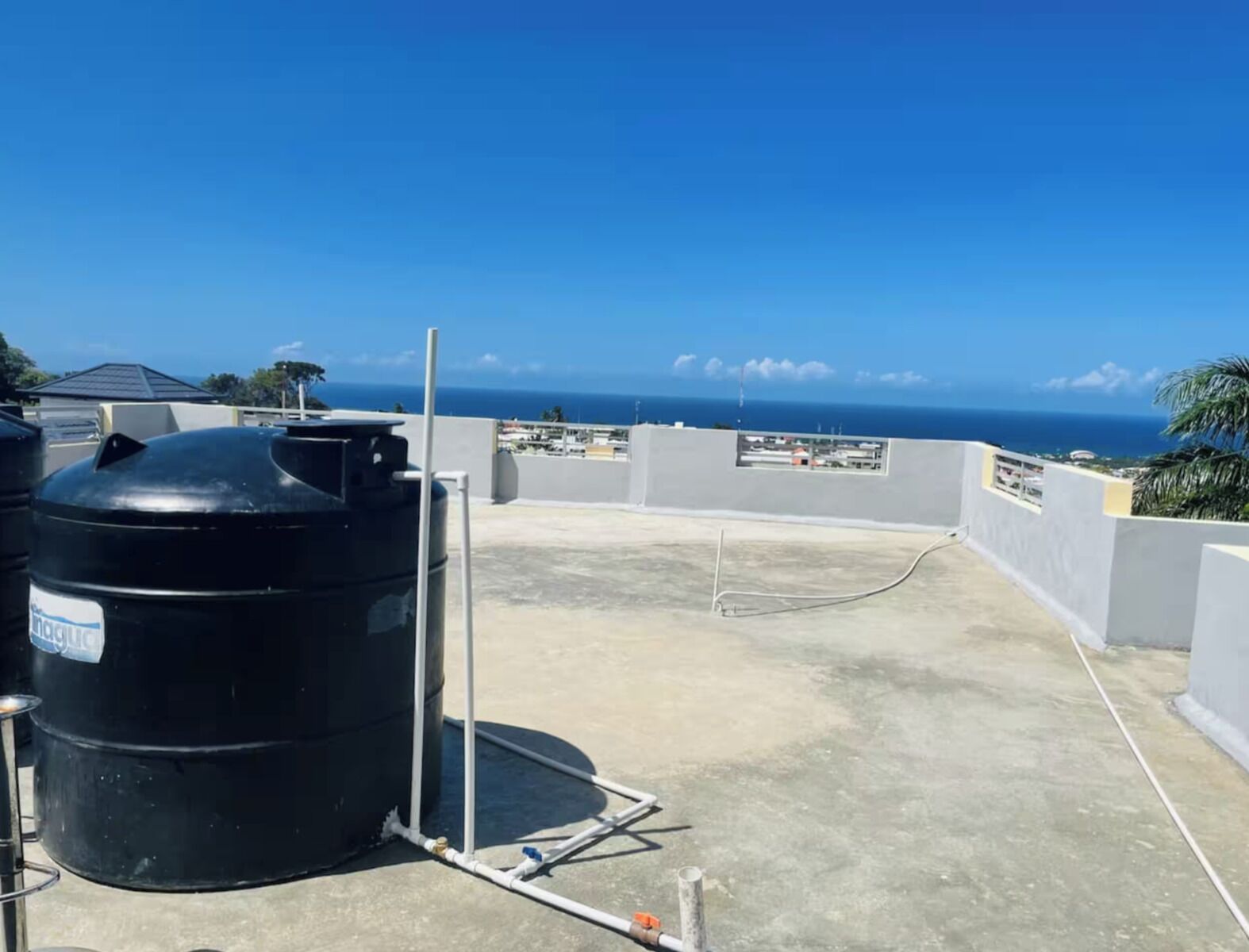Un toit sur lequel se trouve un grand réservoir d’eau noir, des tuyaux blancs et d’où l’on a une vue dégagée sur l’océan.