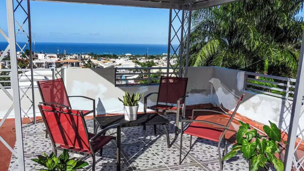 A covered outdoor seating area with chairs and a table, potted plants, and a view of the ocean and cityscape. - 2BR Apartment with Ocean & Mountain Views (Puerto Plata)