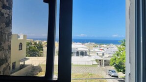 A view through a window with a stone wall on the left, a clear blue sky, and a distant view of buildings and the ocean. - 2BR Apartment with Ocean & Mountain Views (Puerto Plata)
