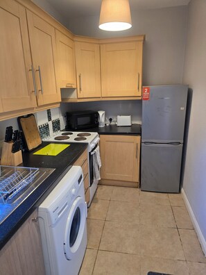 A compact kitchen with wooden cabinets, a stainless steel refrigerator, a microwave, a toaster, and a washing machine.