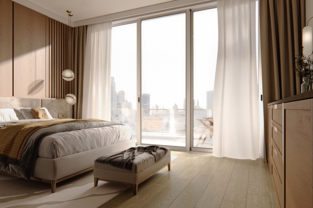 A modern bedroom with a large bed, a bench, a city view, and a wooden wardrobe.