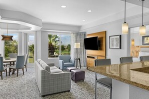Una habitación de hotel moderna con comedor, sofá, televisor y ventanas amplias.