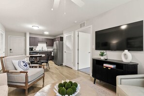 Un salon moderne avec un canapé, une table basse ornée de boules décoratives, un téléviseur fixé au mur et une cuisine en arrière-plan.