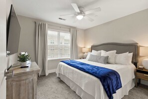 Um quarto com cama, mesas de cabeceira, ventilador de teto, janela com cortinas e TV fixada na parede.