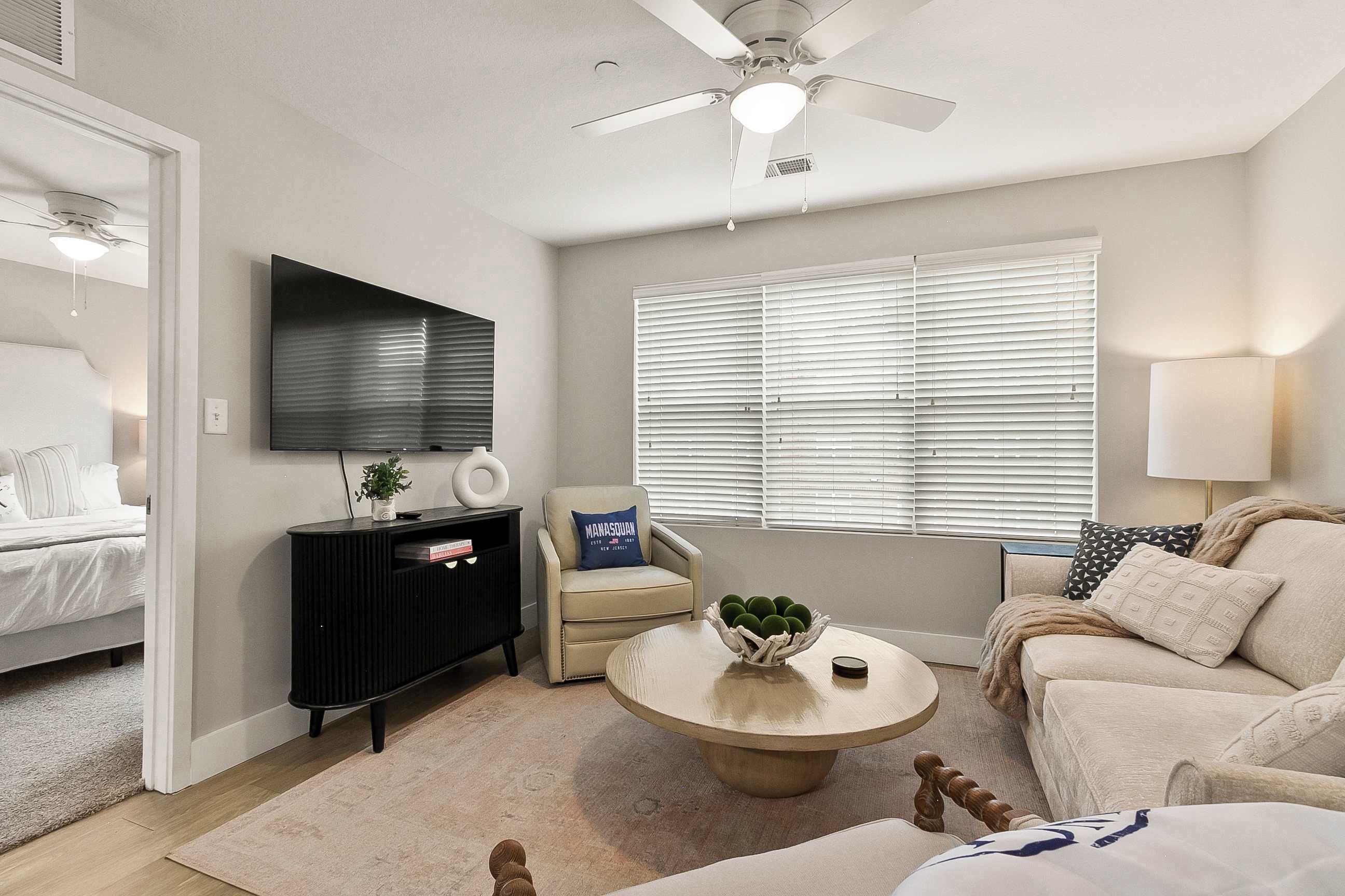 A modern living room with a flat-screen TV, a ceiling fan, a sofa, a coffee table, and a window with blinds.