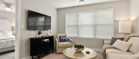 A modern living room with a flat-screen TV, a ceiling fan, a sofa, a coffee table, and a window with blinds.