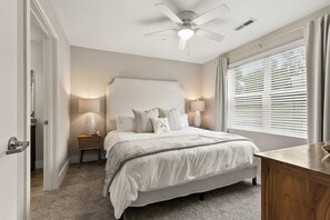 Une chambre à coucher avec un lit, des lampes de chevet, un ventilateur de plafond, une fenêtre avec des stores et un bureau en bois.