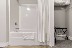 Uma casa de banho com uma banheira branca, uma toalha branca, um chuveiro com cortina branca e uma cesto de roupa branca.