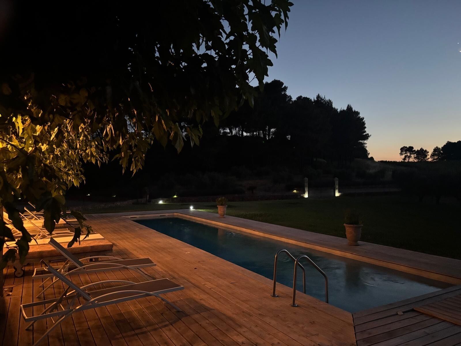 A rectangular swimming pool with a wooden deck, metal loungers, and a small set of stairs leading into the water.