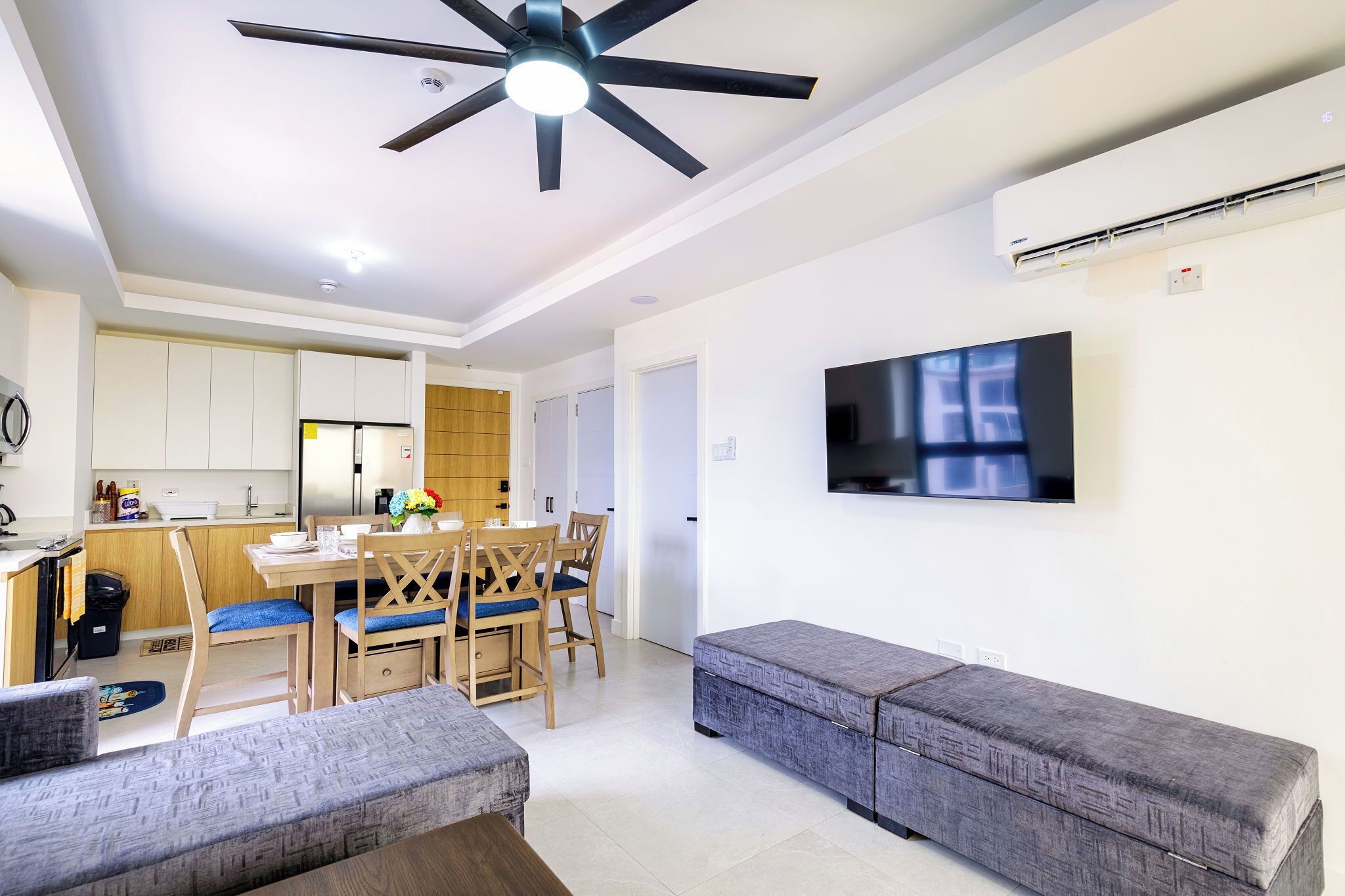 A modern living room with a sofa, a dining table with chairs, a ceiling fan, and a flat-screen TV mounted on the wall.