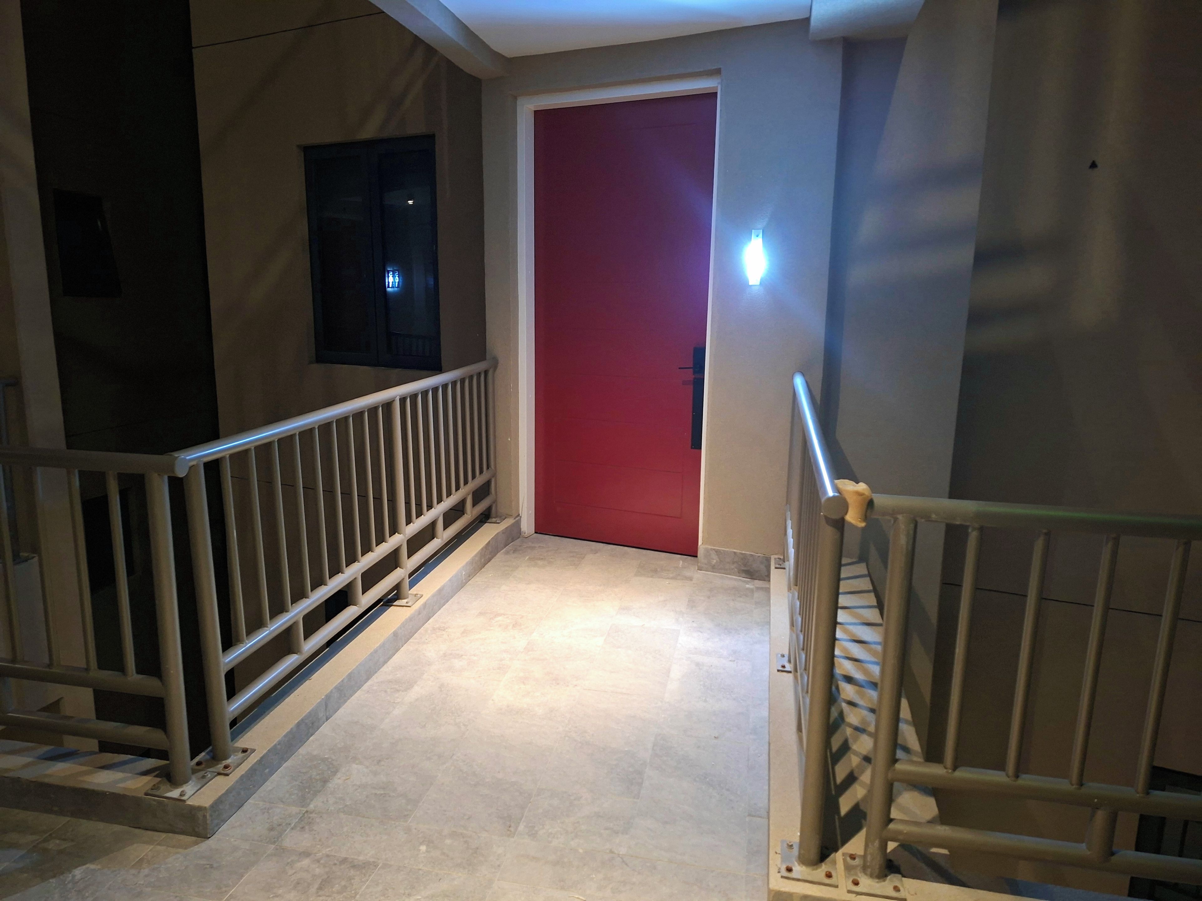 A hallway with a red door, a metal railing, and a light fixture on the wall.
