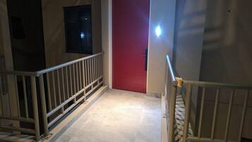 A hallway with a red door, a metal railing, and a light fixture on the wall.