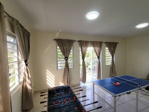 Ein Zimmer mit Kicker, Tischtennisplatte und fensterbelaubten Fenstern.