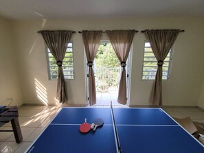 A ping pong table with a net, two paddles, and a ball in a room with windows and curtains. - The Villa in Miradero (Mayaguez)