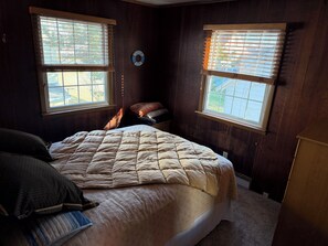 A bedroom with a bed, two windows, a wall clock, and a small table.
