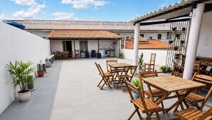 Un patio exterior con mesas y sillas de madera, plantas en macetas y suelo de baldosas.