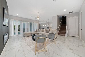 Ein großzügiger Wohnbereich mit einer großen Couch, einem Couchtisch und zwei Sesseln. Eine Treppe führt zu einer weiteren Ebene, und rechts ist eine Tür.