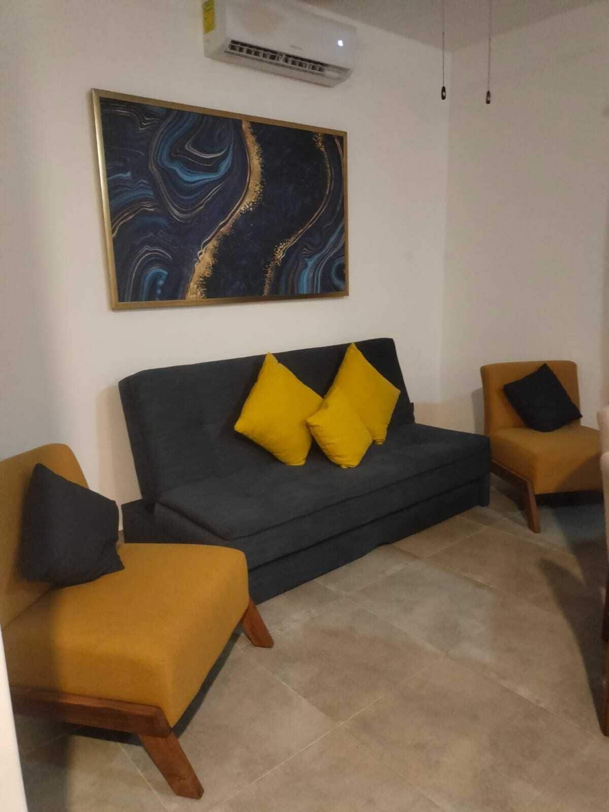 Un salon avec un canapé foncé, deux fauteuils jaunes et un grand tableau abstrait accroché au mur.