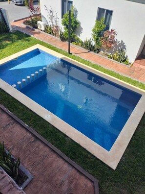 A rectangular swimming pool with a clear blue water surface, surrounded by a well-maintained lawn and various plants. - Family Condo with Pool | Near Beach & 5th Ave | Sleeps 6-8 (Playa del Carmen)