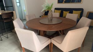 Een ronde eettafel met zes stoelen, een vaas met groene bladeren, en een bank met gele kussens op de achtergrond.