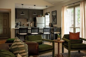 Eine moderne Küche mit dunklen Schränken, einem Kühlschrank und Pendelleuchten. Es gibt eine Essecke mit Stühlen und einem Tisch, sowie ein Wohnzimmer mit grünen Sofas und einem kleinen runden Tisch.