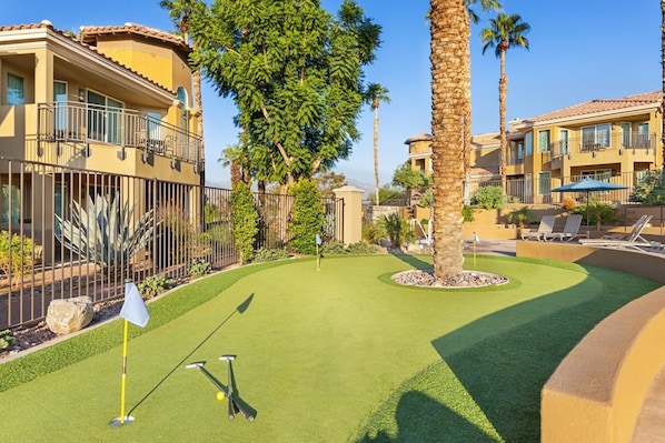 A mini-golf course with a flagstick, golf balls, and a putting cup, surrounded by palm trees and a building with balconies.