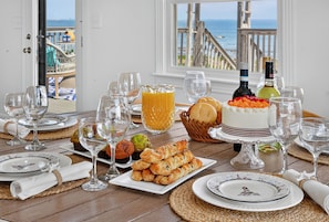 A dining table set with plates, glasses, a cake, pastries, and a bottle of wine, with a view of the beach and ocean. - Oceanfront w/Views, Sleeps 11, Hot Tub, New Reno (Scituate)