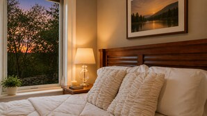 Una cama con cubrecama acolchado, una lámpara de noche, un cuadro con paisaje, y una ventana que da a vista de árboles y un atardecer.