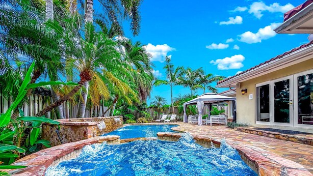 Ein Schwimmbecken mit Steinrand, umgeben von Palmen und einer überdachten Terrasse.