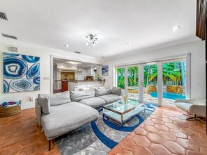 A modern living room with a sectional sofa, a glass coffee table, and a view of a pool through large sliding doors. - Heated Pool & Hot Tub! Pool Table+Close To Beach! (Fort Lauderdale)