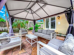An outdoor patio with a covered seating area, stone flooring, and a view of the backyard. - Heated Pool & Hot Tub! Pool Table+Close To Beach! (Fort Lauderdale)