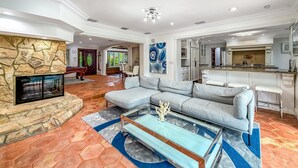 Ein geräumiges Wohnzimmer mit einem Kamin aus Stein, einem großen Ecksofa, einem Glastisch und einem durch einen offenen Bogen einsehbaren Küchenbereich.