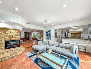 A spacious living room with a stone fireplace, a large sectional sofa, a glass coffee table, and a kitchen area visible through an open archway. - Heated Pool & Hot Tub! Pool Table+Close To Beach! (Fort Lauderdale)